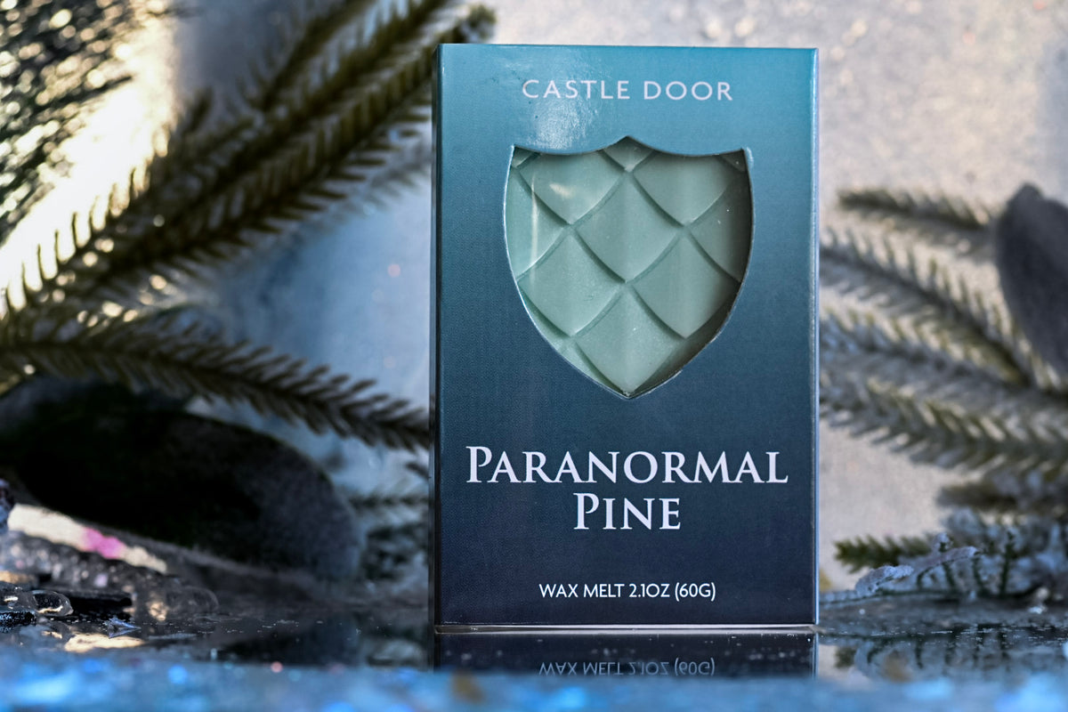 Paranormal Pine wax melt box by Castle Door with pale green dragonscale wax visible through a shield window, surrounded by evergreen branches and snow.