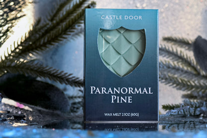 Paranormal Pine wax melt box by Castle Door with pale green dragonscale wax visible through a shield window, surrounded by evergreen branches and snow.