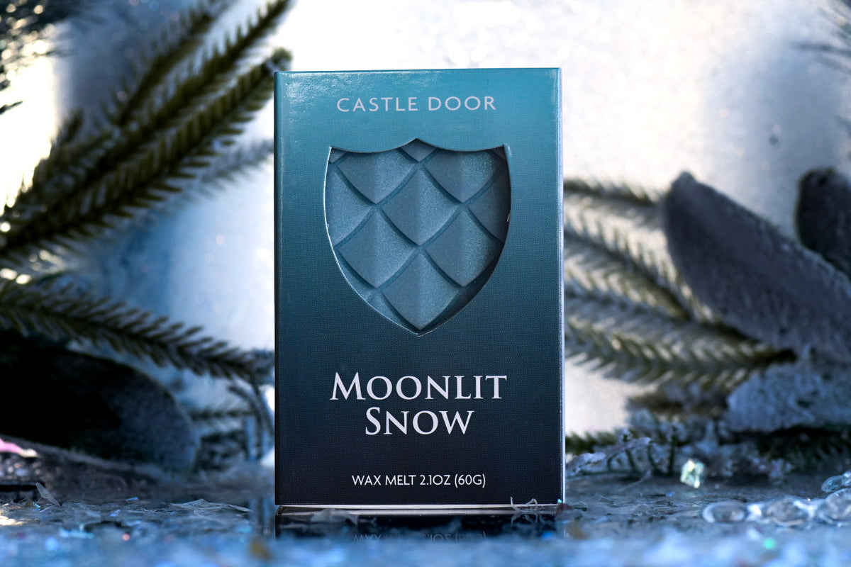 Moonlit Snow wax melt box by Castle Door with icy blue dragonscale wax visible through a shield window in a snowy winter setting.